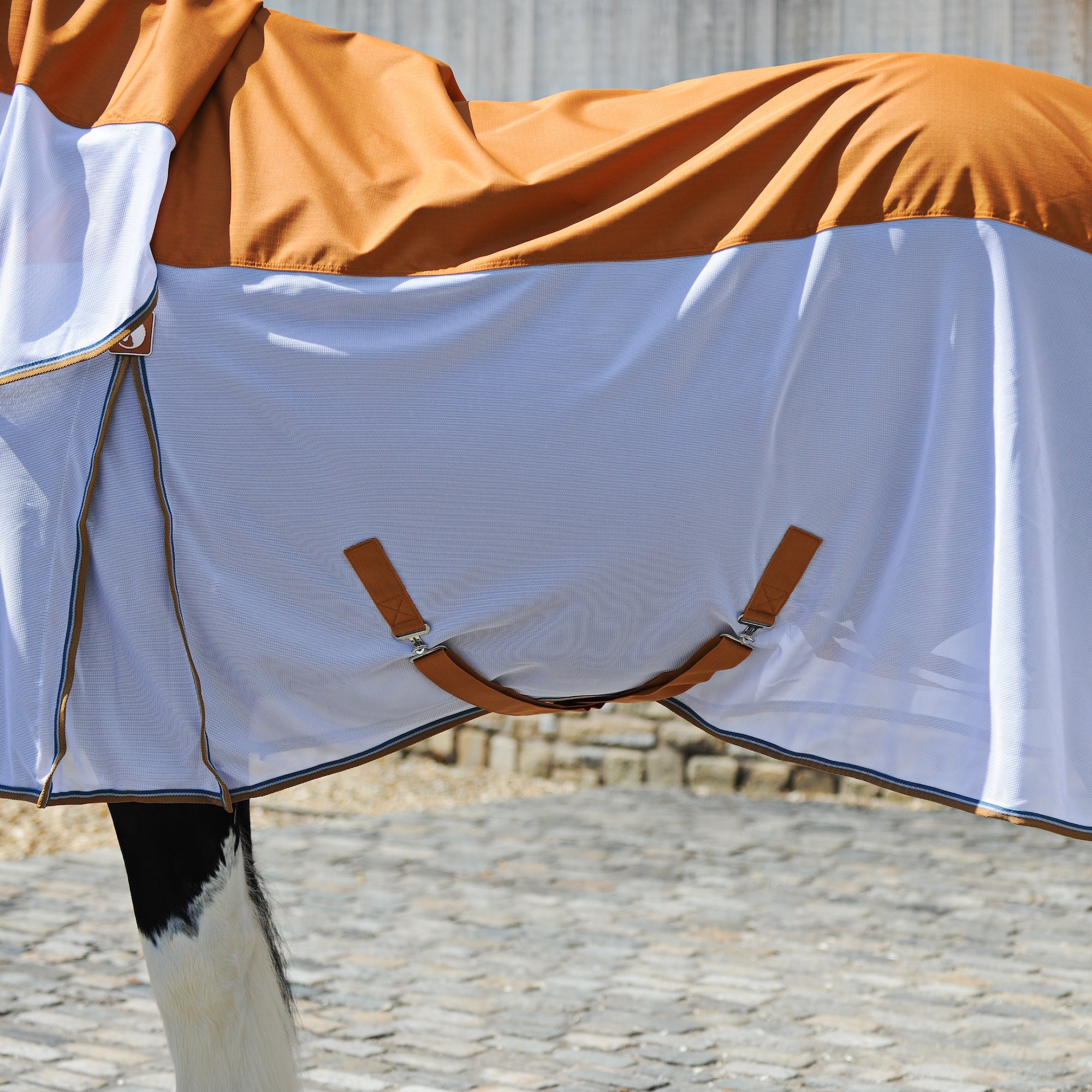 A horse turnout blanket with a white mesh lower section and a warm orange waterproof upper section, draped over a horse standing on a cobblestone surface.