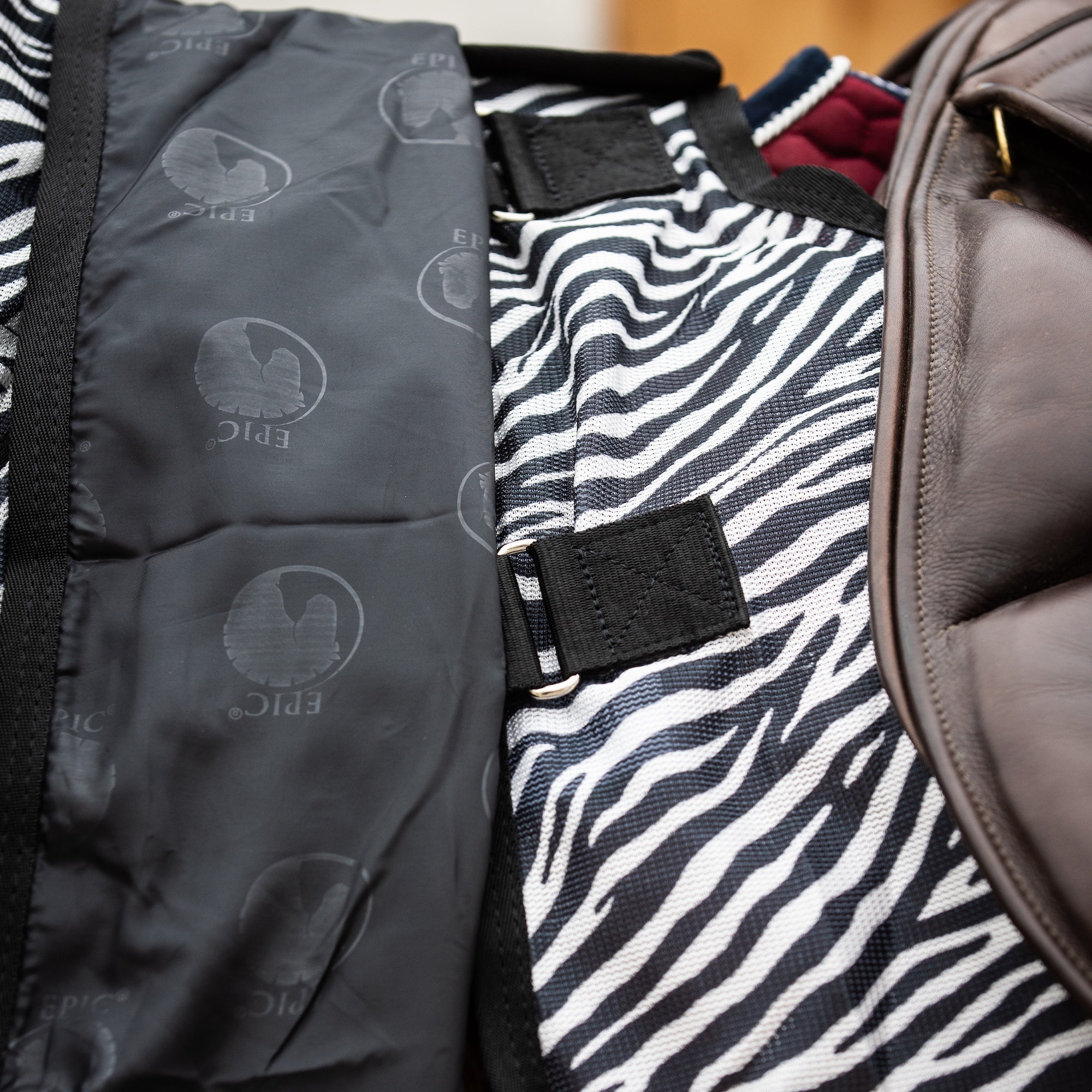 Close-up of a ride-on horse fly rug showing a black and white zebra-print fabric panel, a soft black lining with subtle logo print, and a velcro attachments.