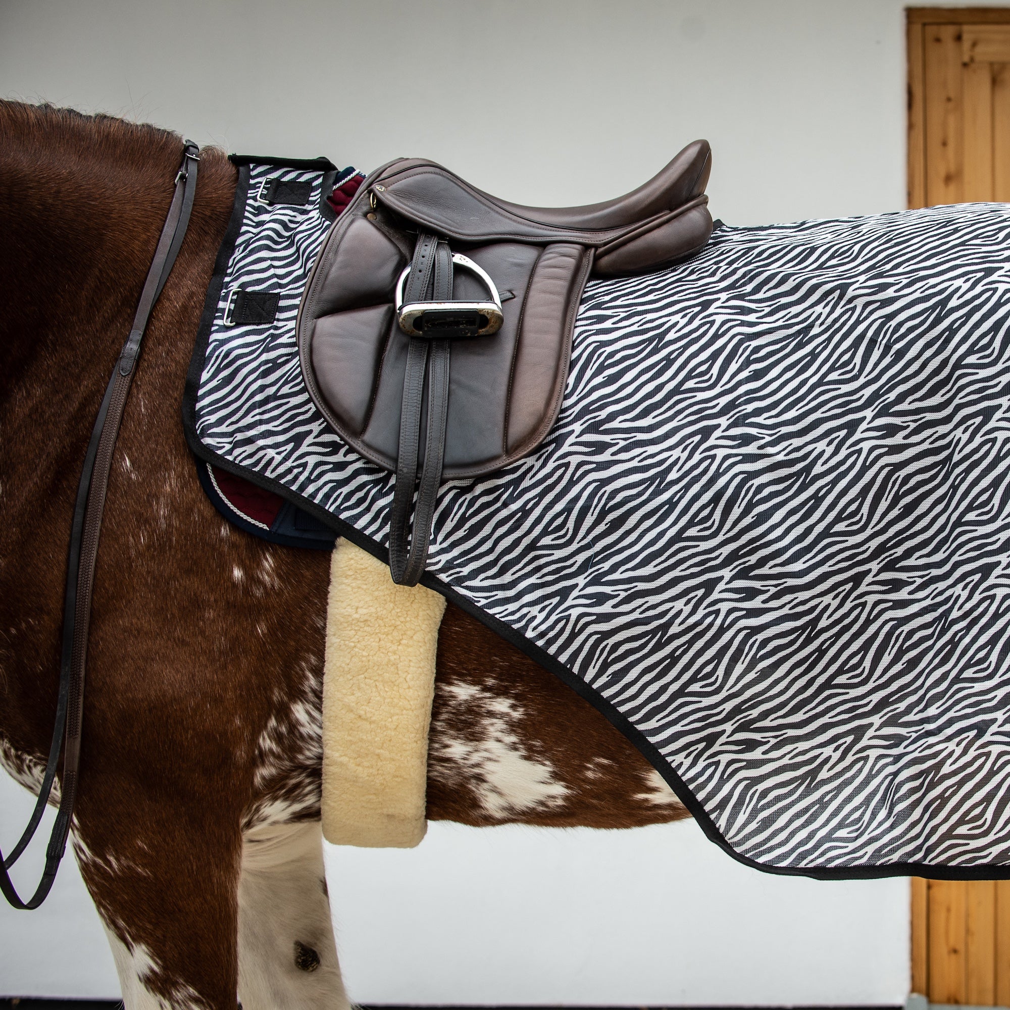 A horse blanket with a black and white zebra pattern and a beige fleece lining, shown on a brown and white spotted horse with a dark brown leather saddle.