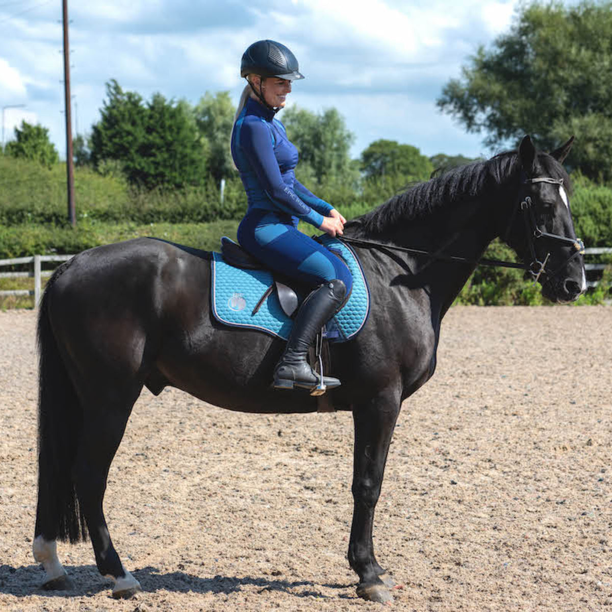 A person riding a black horse in an outdoor arena, sitting on a blue saddle pad which is the product in question, appears to be of average height and dressed in equestrian attire including a helmet, long-sleeved top, and riding pants.