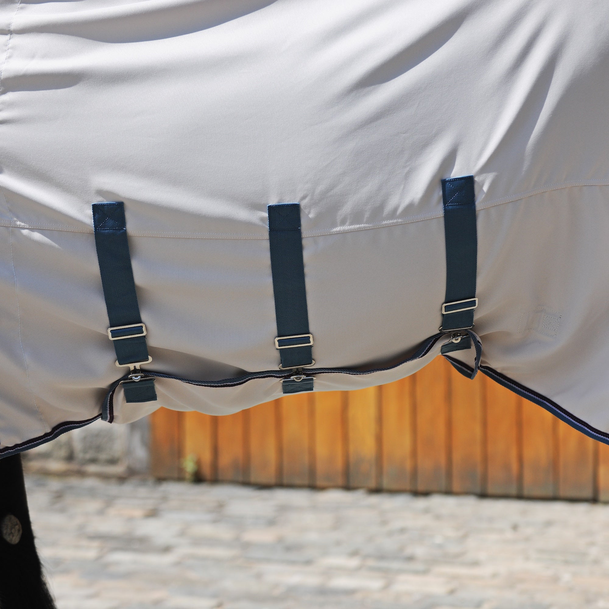 Close-up of a white horse blanket with dark navy straps and silver buckles, shown outdoors against a blurred wooden fence and stone ground.