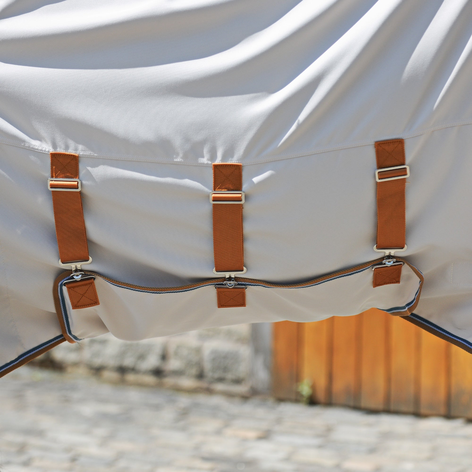 Close-up of a white belly piece on a sweet itch rug with three brown strap attachments and a matching brown trim, photographed outdoors with a blurred stone wall and wooden fence in the background.