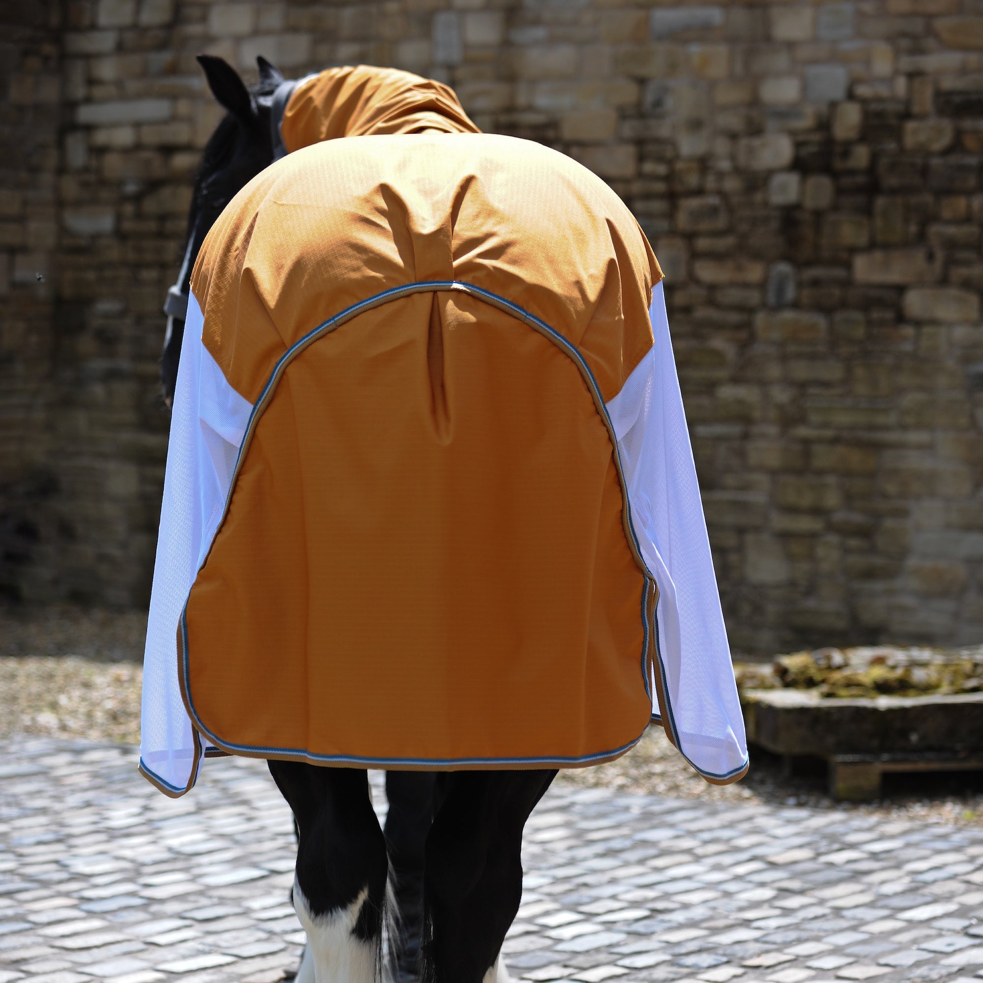 A horse wearing a orange and white fly blanket with a hood, photographed from behind in an outdoor stable setting with a stone wall and cobblestone floor.