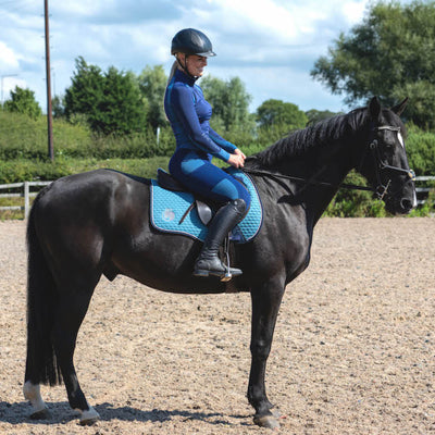 A person riding a black horse in an outdoor arena, sitting on a blue saddle pad which is the product in question, appears to be of average height and dressed in equestrian attire including a helmet, long-sleeved top, and riding pants.